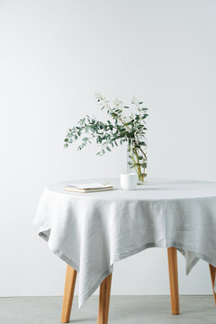 Side View Round Table With A Rough White Tablecloth And Branch In A Jar. Cropped, Over White Wall. Indoors. Notepad And Cup On A Surface.