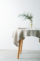 Low angle image of a served round table with a white tablecloth. Empty glass, unlit candles and decorative branch. Over a white wall.