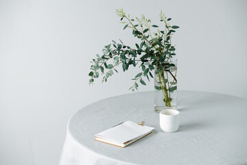 Notepad on a round table with a rough tablecloth and branch in a jar. Cropped, over white wall....