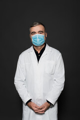 senior physician in white coat and medical mask looking at camera isolated on dark grey.
