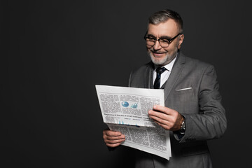 happy senior man in suit and eyeglasses reading newspaper isolated on dark grey.