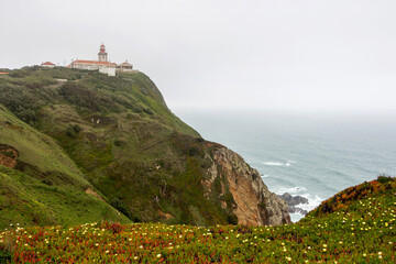 Lighthouse on a hill in cloudy weather at Cape Roca in Portugal, greenery with yellow flowers, sheer brown cliffs with stones and waves in the turquoise water below