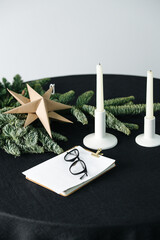 Carton origami star, small artificial tree, notepad, glasses and unlit candles on a round table with a black rough tablecloth. Cropped