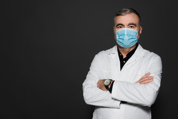 doctor in white coat and medical mask standing with crossed arms and looking at camera isolated on dark grey.