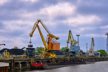 Fototapeta na wymiar River port cranes with clamshell or griper loading coal to river drag boats or barges moored by pier on cloudy day. Energy, minerals, environment 