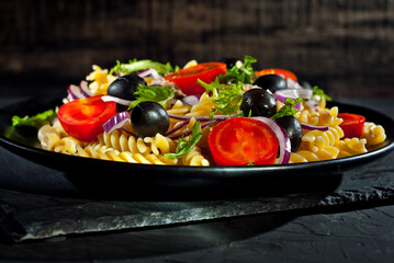 Salad with tomatoes, peppers, tuna olives and pasta. Fresh salad in a black plate on a dark background.