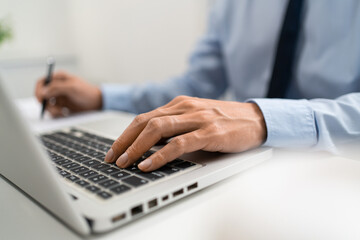 Obraz premium Working Man Conept The male officer sitting in the office, laying his left hand on the laptop, and writing something on the paper with his right hand