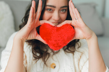 Emotional portrait of 30 year old woman in knitted stylish cardigan. Young beautiful dark haired curly woman holding red plush card heart on valentine's day in her hands.