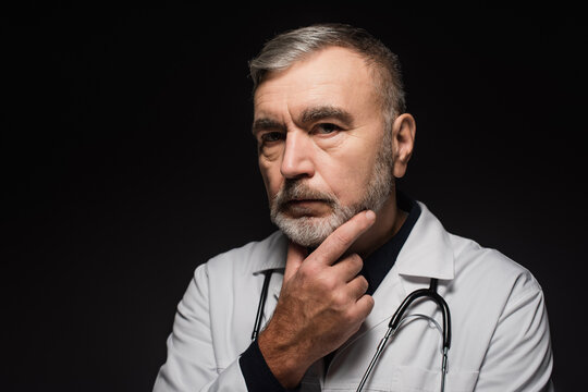 pensive bearded doctor looking at camera while touching chest isolated on black.