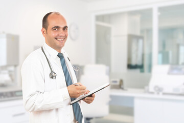 Male doctor with stethoscope holding a notebook. Smiling male doctor filling out medical form on clipboard while standing straight in hospital