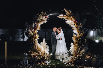 A stylish groom and a beautiful bride in a long dress are standing at night near a reed arch decorated with lamps and garlands. Wedding photography, portrait. - obrazy, fototapety, plakaty