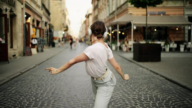 Funky tattooed woman in wireless headphones dancing alone in the street. Beautiful sexy woman with tattoo dancing in downtown very emotional. Attractive girl wearing blank white t-shirt and jeans