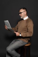 stylish senior man in eyeglasses reading newspaper while sitting on stool on dark grey.