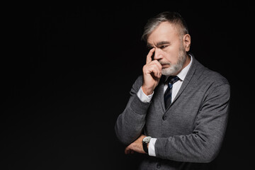 pensive senior man in grey jumper touching face while thinking isolated on black.