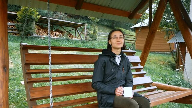 Young attractive farmer woman drinking coffee while sitting on a wooden swing.A young attractive woman is drinking coffee and enjoying a beautiful view of the mountains and the field. 