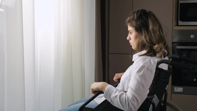 Pensive disabled woman with infantile cerebral paralysis sits in wheelchair near window behind curtain at home experiencing ableism side view.
