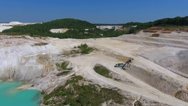 Uneven territory of kaolin clay mine. White landscape with excavator loading the truck machine. Green trees and blue clear sky backdrop.
