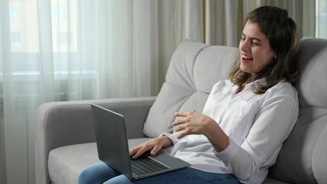 Young disabled woman with cerebral palsy talks gesticulating at job interview via camera on laptop sitting on sofa near window in living room at home.