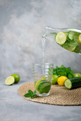 Pouring tasty fresh cucumber water from bottle into glass. A bottle of clean, cool and fresh drink stands on a gray concrete background with ingredients on board. Strengthening immunity, diet.