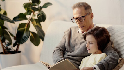 middle aged grandfather in glasses reading book to grandchild at home