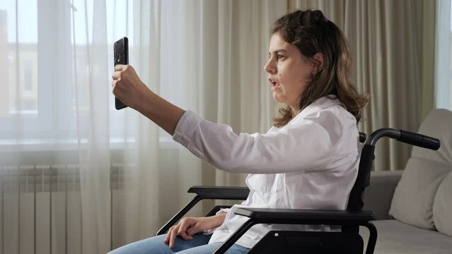 Cheerful disabled woman with organic damage of central nervous system talks emotionally on videocall via phone sitting in wheelchair at home side view.