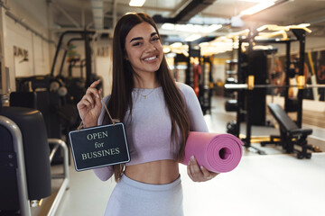 A beautiful young girl, looking at the camera with a smile on her face, holds a sign that the gym has reopened after a long time. A young woman holding a yoga mat indicates that the gym has reopened.