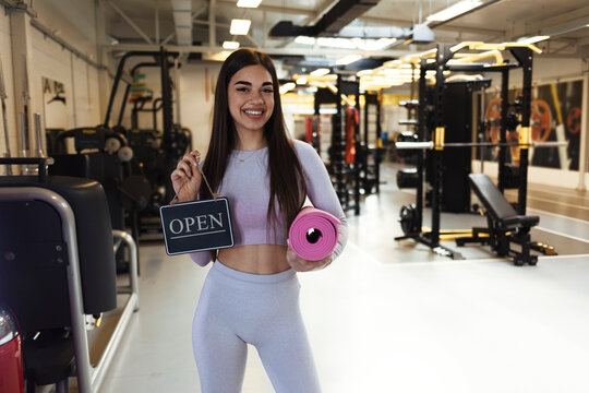 Portrait of smiling trendy 20 years old gym owner woman, holding mats and showing open sign. Opening fitness centar after covid-19 pandemic. People and healthcare concept. Copy space.