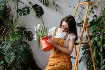 Young female gardener wearing denim and eyeglasses overalls taking care about houseplants at home garden, woman florist sitting on ladder with orchid flower in milk can, growing indoor plants as hobby