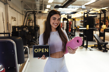 Obraz premium A beautiful young girl, looking at the camera with a smile on her face, holds a sign that the gym has reopened after a long time. A young woman holding a yoga mat indicates that the gym has reopened.
