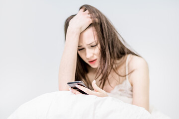 Fototapeta premium A pretty brunette holding her head with a sad look sits in bed, covered with a blanket, holds a phone in her hands, looks at it and types. The girl has bad news. Good morning