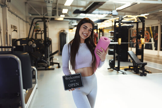Cropped shot of a gym instructor holding up a "open sign". Attractive young woman opening up her fitness center. Portrait of a sporty young woman holding an exercise mat and "open" sign in a gym.