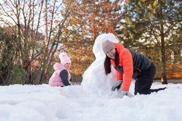 Portrait of small girl and mother crawling on snow near snowman on backyard in evening with rowan and fir trees in background. Parents spending time with children.