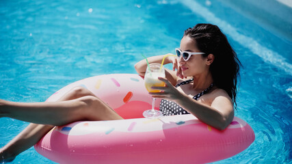 cheerful woman in sunglasses holding glass with cocktail and swimming on inflatable ring in pool