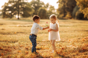Fototapeta premium A boy in a white shirt and blue jeans gives a girl with blond hair in a white dress a flower. They stand in a field at sunset with a beautiful view.