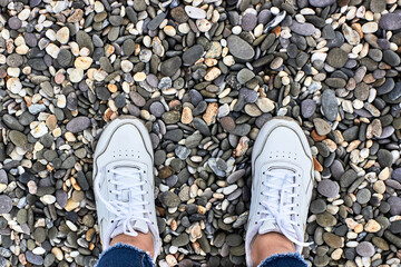 A girl in white sneakers and jeans with fringe is standing on a grey pebble beach. Women's feet on the sea coast, sea stones. Top view.
