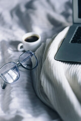 glasses and laptop. a cup of coffee, laptop glasses on the background of a white bed and a light plaid. relaxation and comfort. work from home. sickness work in bed