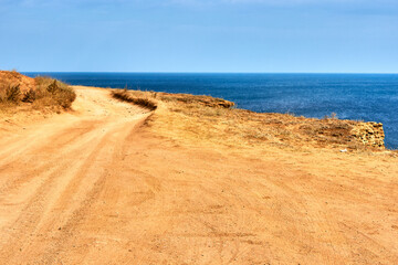 A sandy country road along the edge of the cliff. Orange land, a cape by the sea. Seascape, horizon, calm blue sea, clear sky.