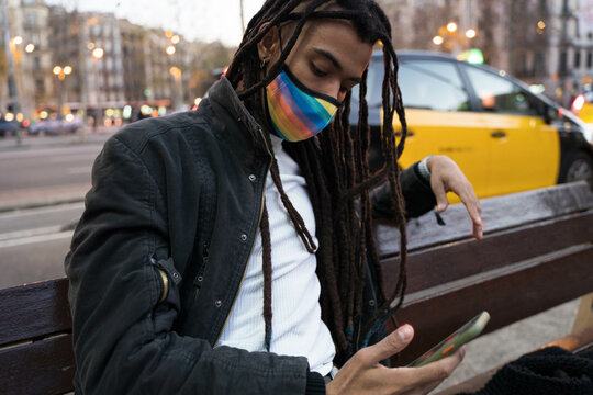African American boy sitting in the city looking at his cell phone with LGTB mask. 