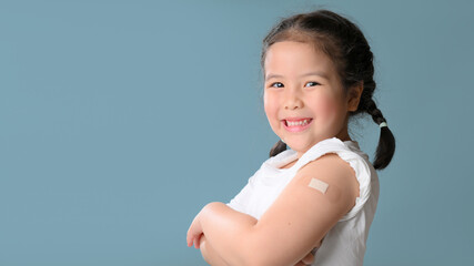 Coronavirus Vaccination Advertisement. Happy Vaccinated Little asian child girl Showing Arm With Plaster Bandage After Covid-19 Vaccine Injection Posing Over Blue Background, Smiling To Camera.