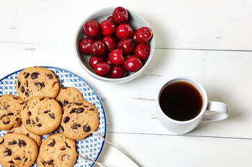 White cup with coffee, traditional cookies on plate and sweet cherries in a ceramic bowl on a white wooden background, closeup. Tasty breakfast, morning routine concept