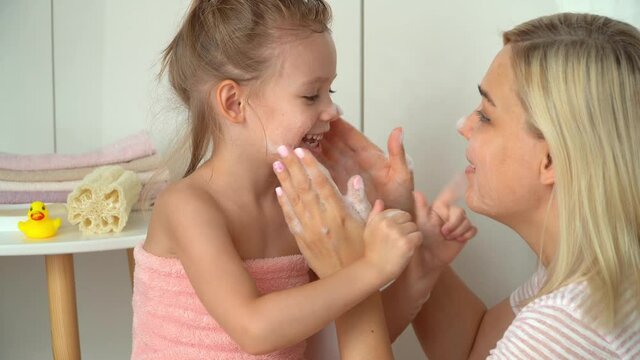 Mother and little daughter fun wash each others face with soapy foam in bathroom. Bathing child with soap, hygiene and health concept. Baby care