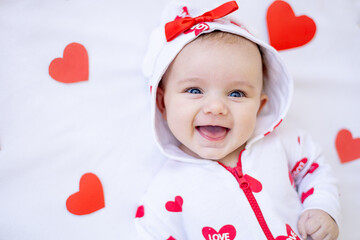 portrait of a funny baby girl with red hearts on a bed on a white cotton bed smiling or laughing, valentine's day concept