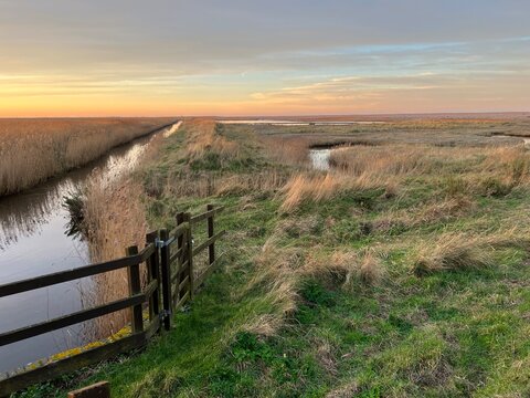 Sunset landscape of Cley next the sea beautiful marshland nature reserve in Norfolk East Anglia uk on the coast with river water, reeds, fencing and colourful sky on horizon by beach in Winter