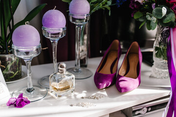 Wedding accessory bride. Stylish violet women shoes, earrings, bouquet flowers, candles and perfumes on table standing on wooden background table.