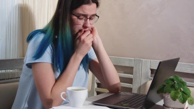 Tired girl student with glasses with blue hair is typing on a laptop, drinking coffee, yawning. Girl falls asleep at the computer