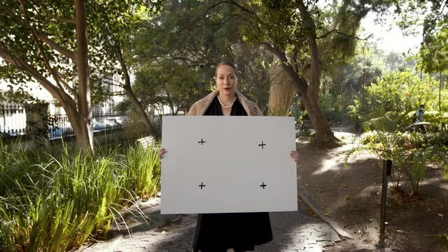 Posh woman holding a blank poster while standing in a park. High class woman displaying a white placard outdoors.