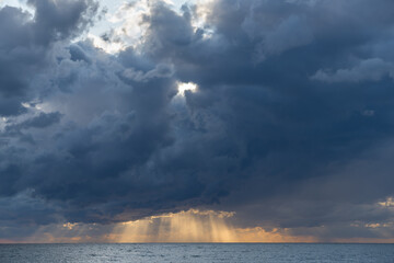 Deep dark blue clouds with rare solar gaps descended low over the Black Sea. The yellow rays of the sun are visible, illuminating the water. The texture of the stormy sky. Volumetric natural photo 