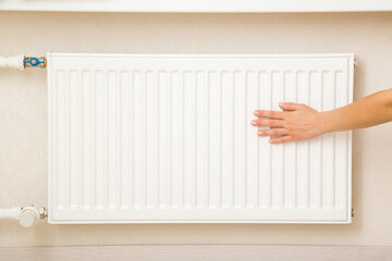 Young adult woman hand touching white heater and checking warmth in room. Heating season. Closeup. Front view.