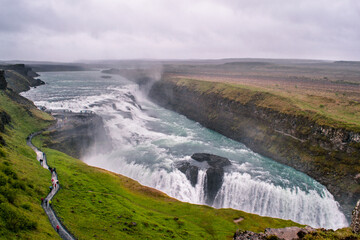 Wideangle panoramic view of Gullfoss Falls with turquoise water on a rainy day. Cloudy sky and black volcanic soil. Tourists' path on the side of the falls. Part of the Golden Circle tour, Iceland