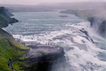 Wideangle panoramic view of Gullfoss Falls with turquoise water on a rainy day. Cloudy sky and black volcanic soil. Tourists' path on the side of the falls. Part of the Golden Circle tour, Iceland